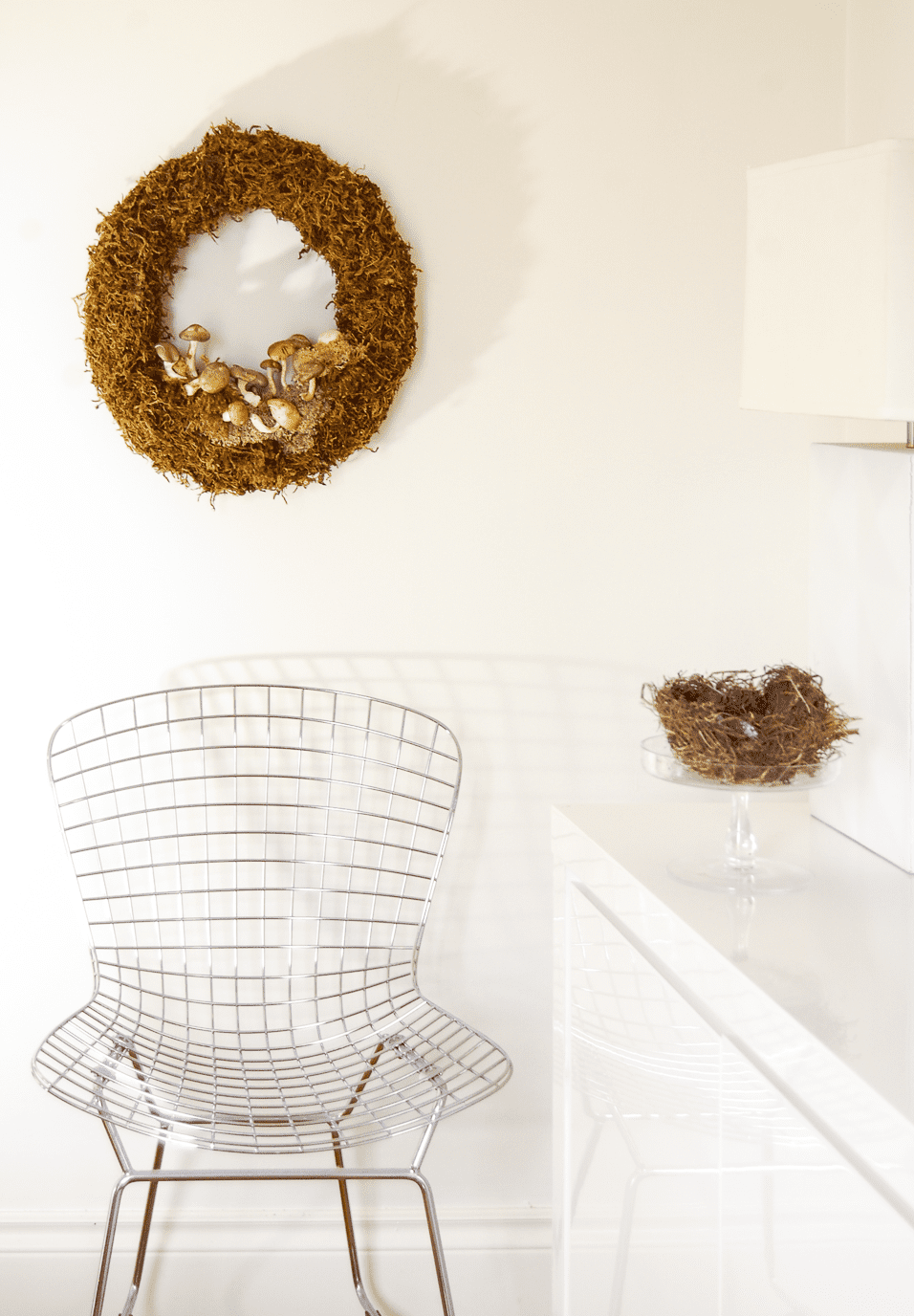 Moss wreath with mushrooms hangs on white wall over Bertoia chair.