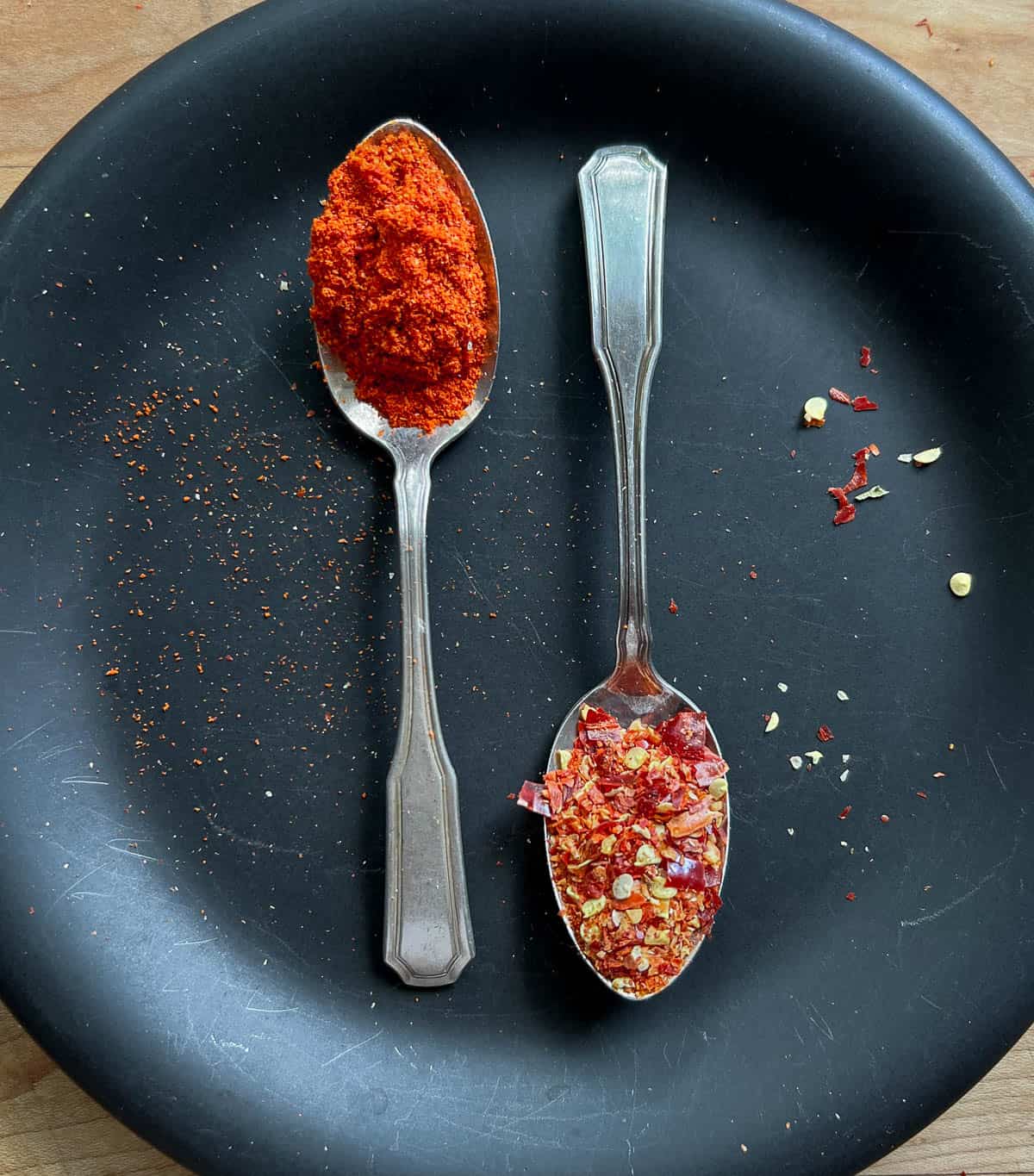 Two silvers spoons on a black plate with cayenne pepper in one and hot pepper flakes in another.