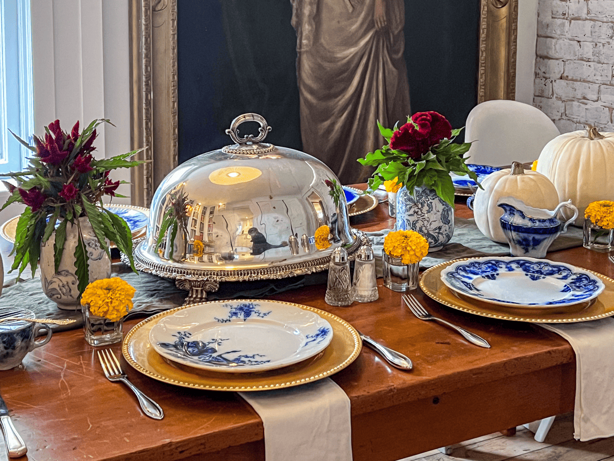 Harvest table set for dinner with gold chargers, silver cloche, and flow blue plates.