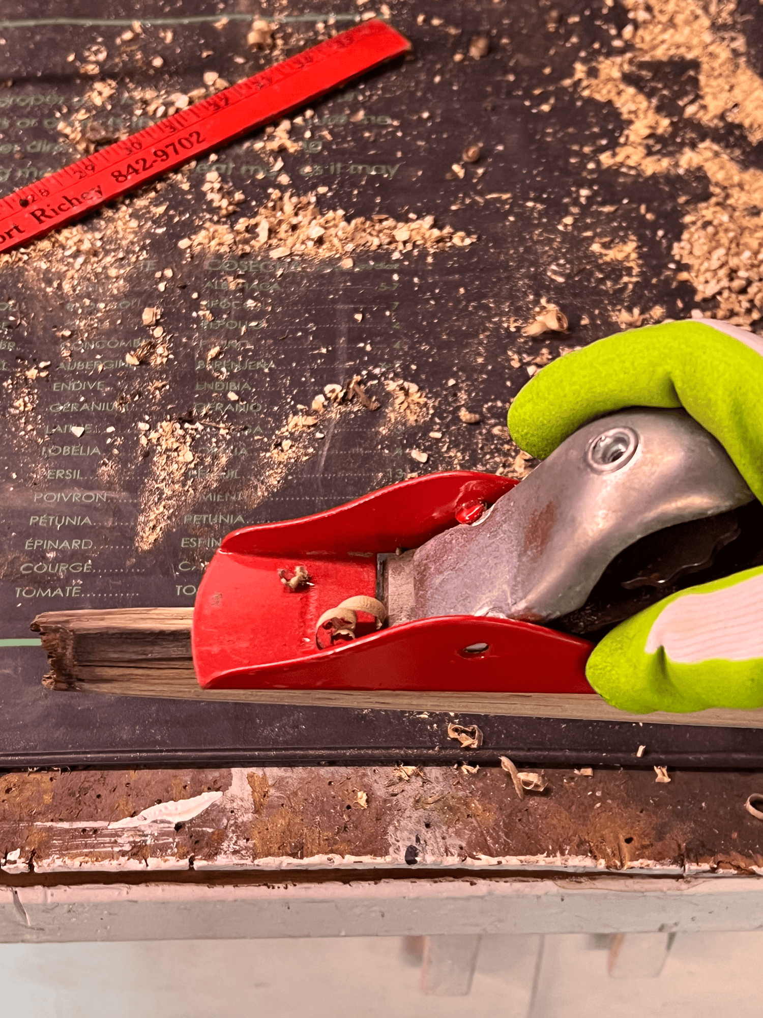 A green gloved hand holds a red hand planer as it shaves off the end of a wood shovel handle to taper it.