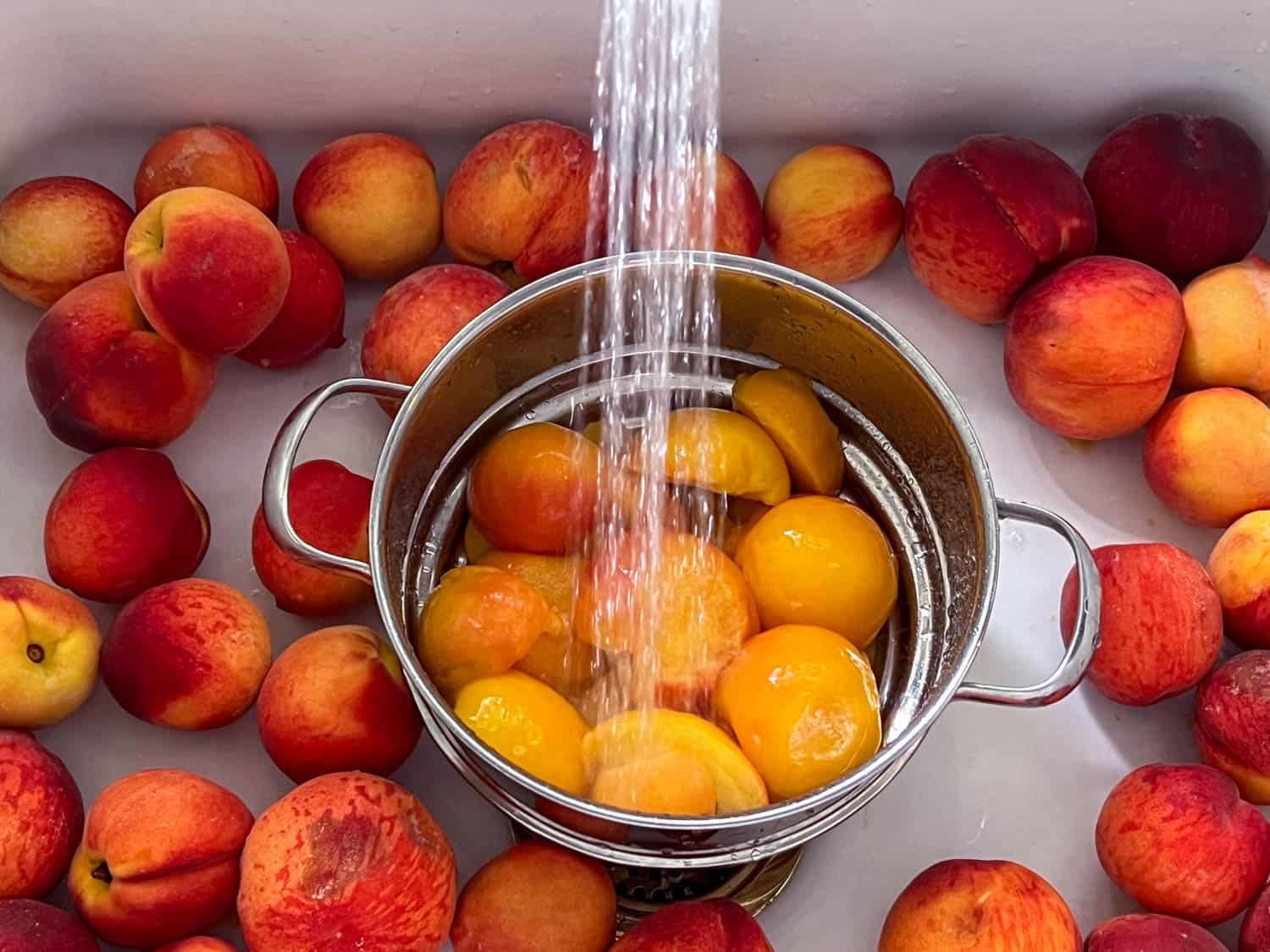 Peeled peaches in a stainless colander being rinsed with water in a white enamel sink.