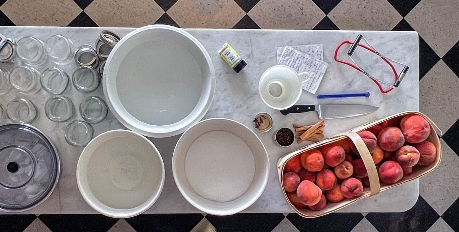 A marble island covered in equipment and ingredients needed for canning peaches including a basket of peaches, bowls of sugar, ice, mason jars, spices and canning tools.
