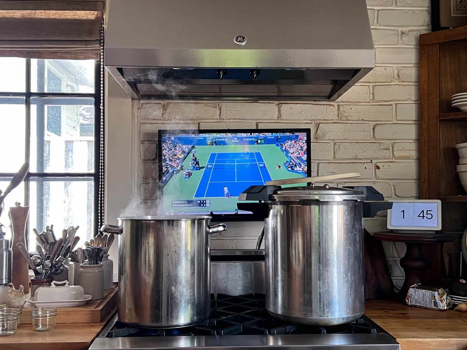 A kitchen scene with 2 large pots on a range, a tv with tennis on it, and a clock that reads 1:45.