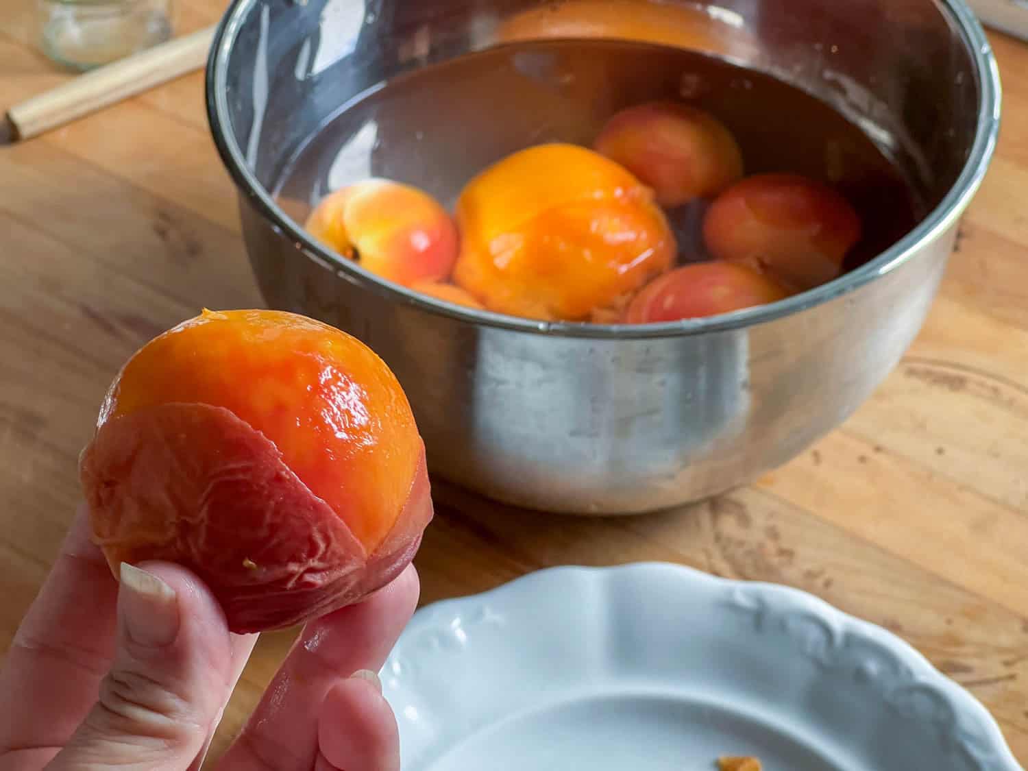 A peach skin being easily pinched off of a peach with a bowl of peaches in ice water behind.