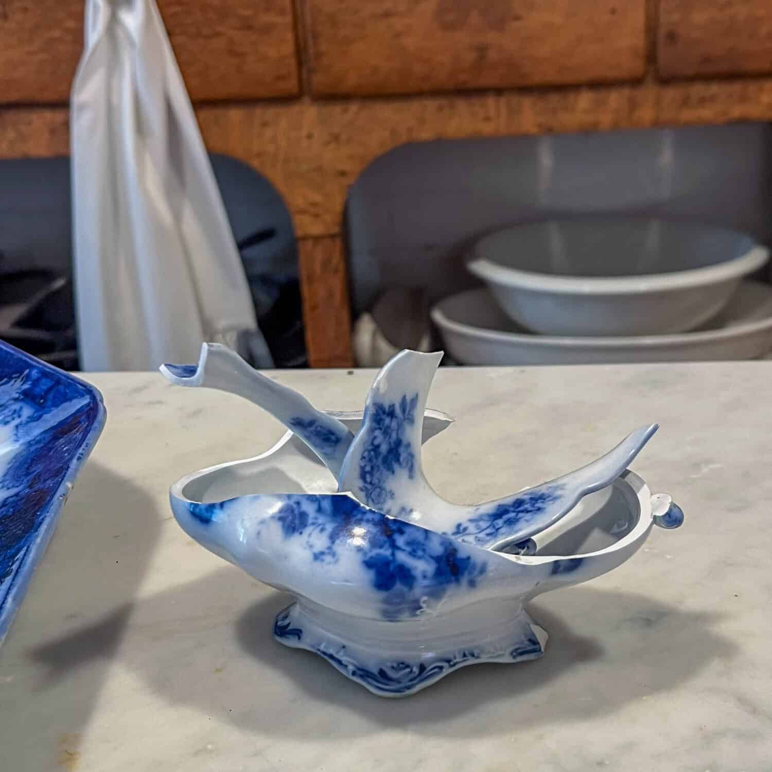 A shattered antique flow blue gravy boat sits on a marble countertop.
