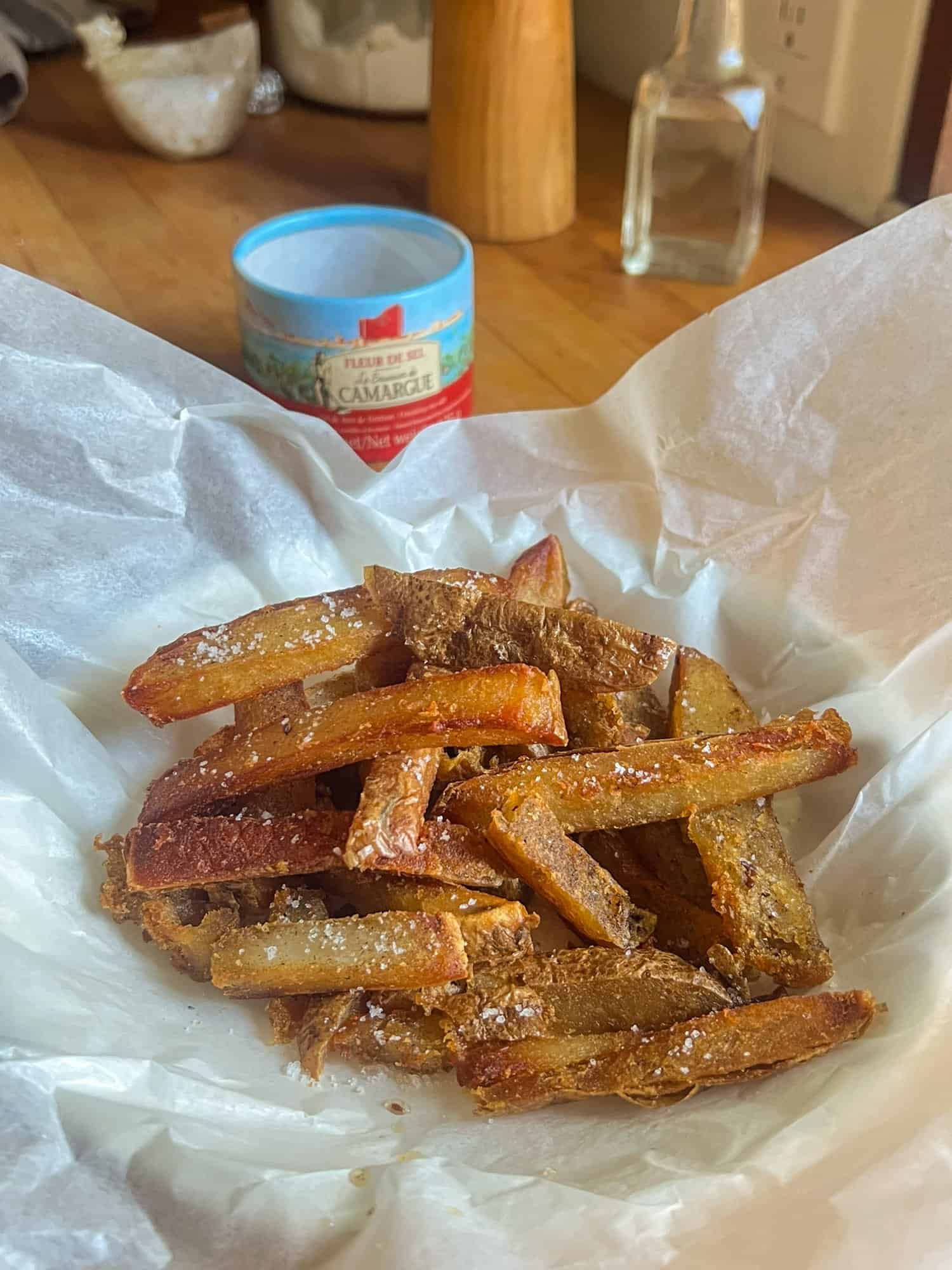 A plate lined with parchment paper filled with salt sprinkled french fries with the skin on made from baked potatoes, salt in the background.
