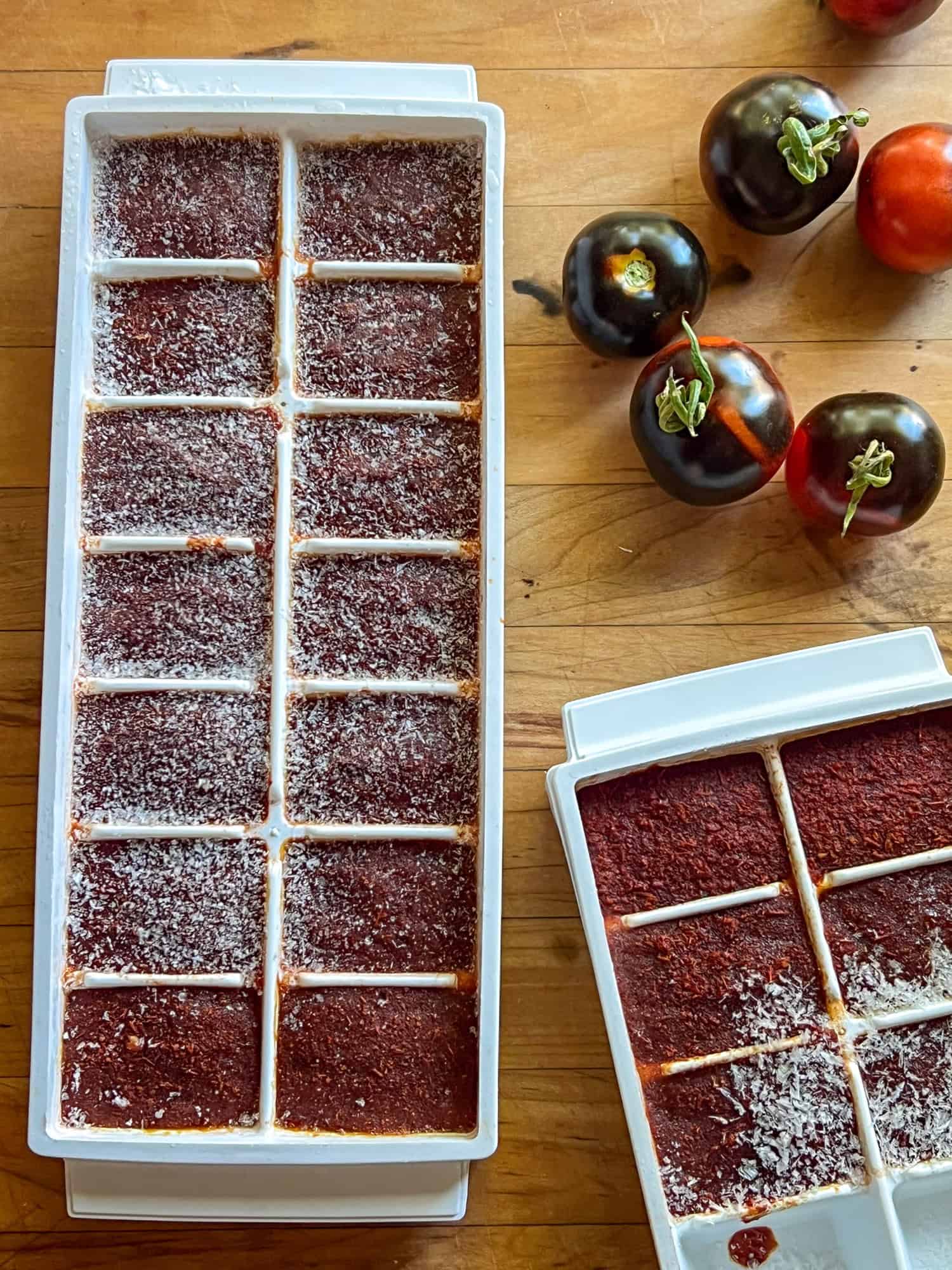 A white ice cube tray filled with frozen dark red tomato sauce and small tomatoes around.