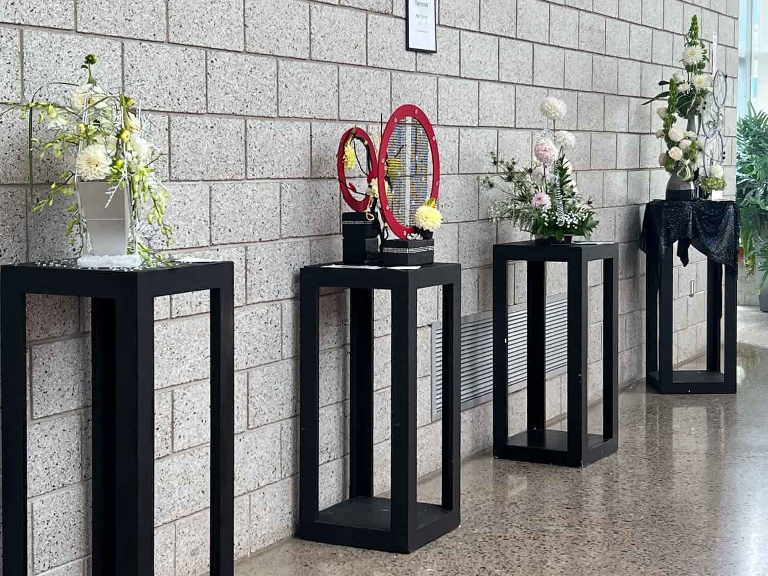 A competition row of exhibition flower arrangements along a cinder block wall.