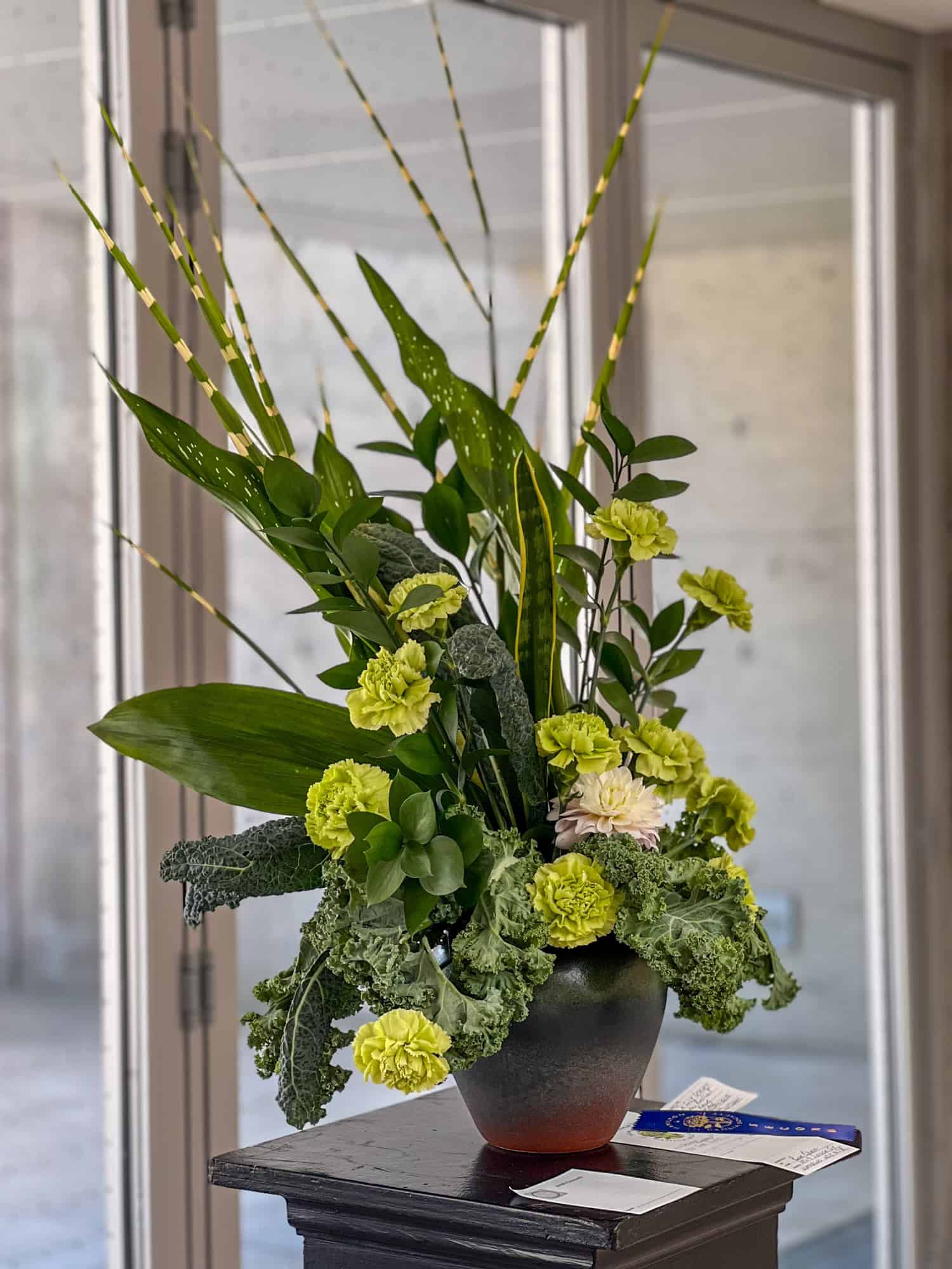 A high reaching floral arrangement in green with carnations, snake plant, and several t ypes of kale.
