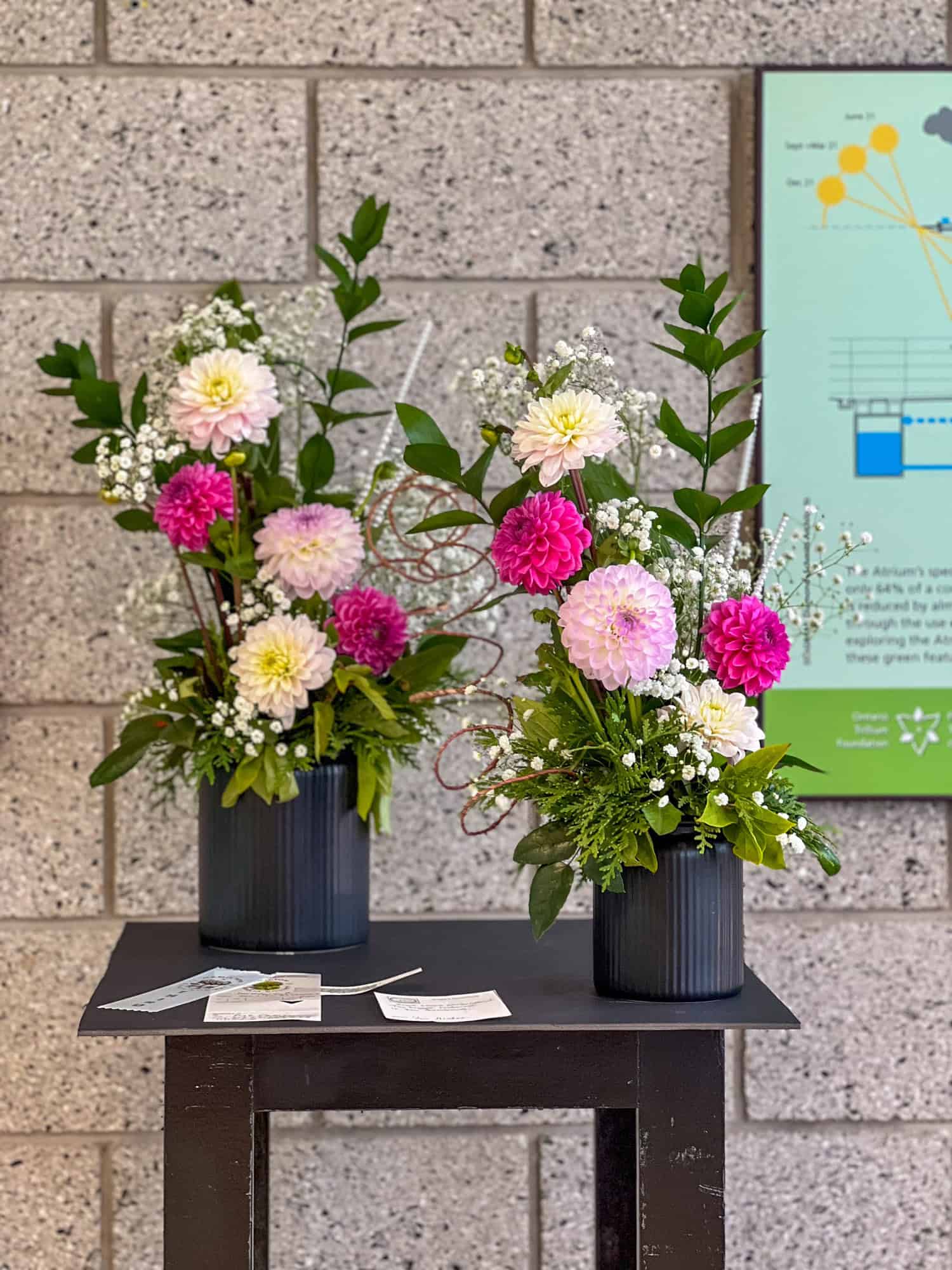 2 exhibition floral arrangements of various pink dahlias with greenery in ceramic black ribbed vases.