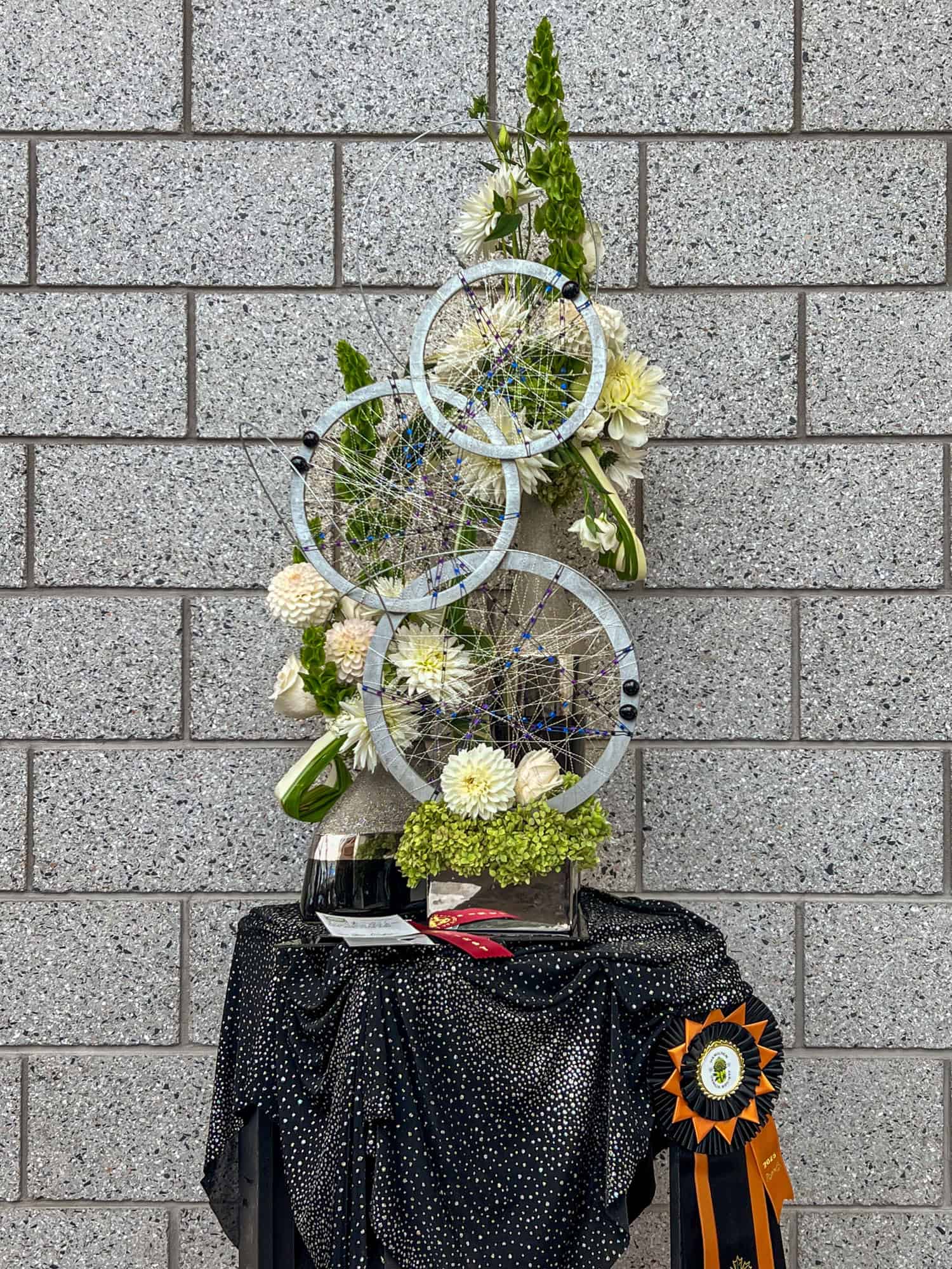 Tall exhibition floral arrangement with round, dreamcatcher type adornments.